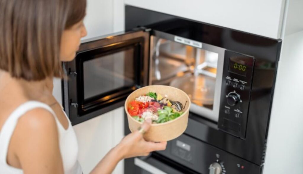 A woman placing an open container of veggies in a microwave oven