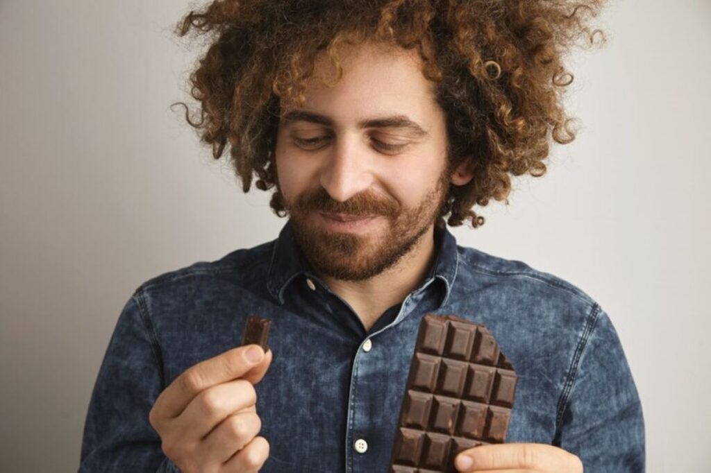 Young happy bearded man with healthy skin and curly hair pleasured with the taste of a chocolate bar