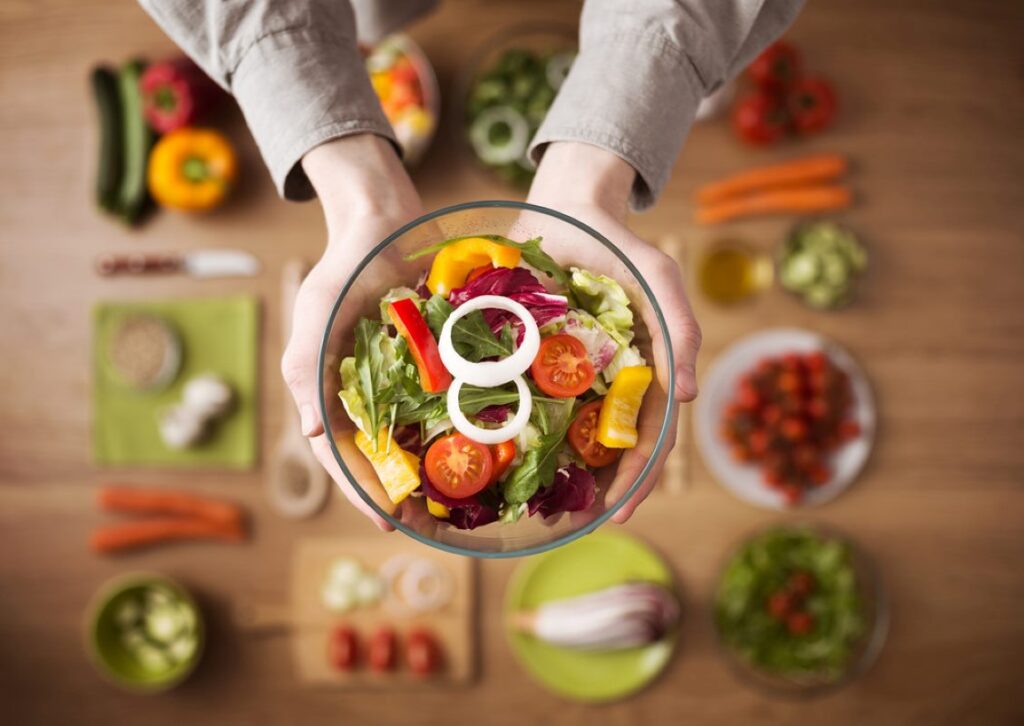 Hands holding an healthy fresh vegetarian salad in a bowl, fresh raw vegetables on background, top view