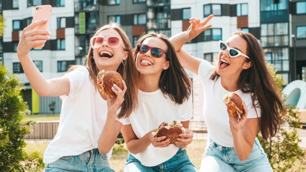 Three young beautiful smiling hipster female in trendy summer clothessexy carefree women posing in the streetpositive models taking selfie in sunglassesholding juicy burger and eating hamburger