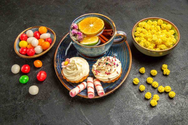 Side view a cup of tea bowls of colorful sweets next to the plate of an appetizing cupcake with cream and a cup of tea on the table