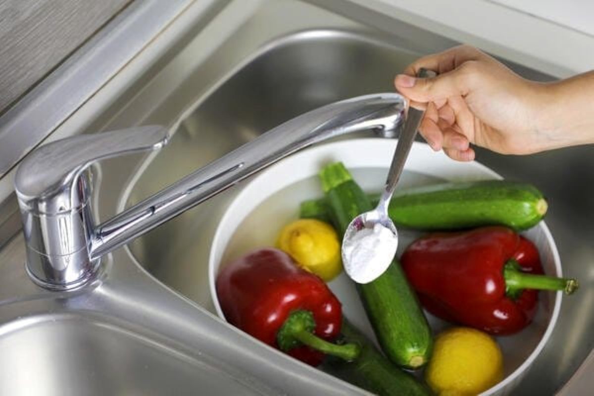 Washing fresh produce in a kitchen sink, with a spoonful of baking soda hovering over the bowl